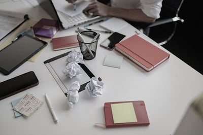 Cluttered white desk with notebooks, phones, sticky notes, crumpled paper, and a tablet, while a person works on a laptop in the background.