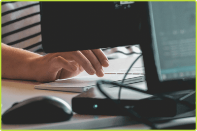 Close-up of hands using a keyboard at a desk, framed by computer monitors, with a mouse and small switch in the foreground.
