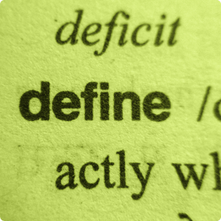 Close-up of printed dictionary text showing the word “define” in bold, on yellow-green paper.