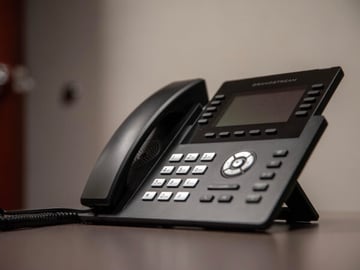 Black Grandstream office desk phone angled on a desk, with handset, keypad, display screen, and blurred neutral background.