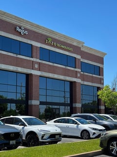 Exterior view of the office building entrance area with Inzo Technologies signage above reflective windows and parked cars in front.
